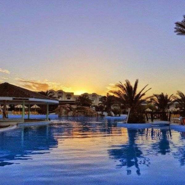 Pool im Djerba Holiday Beach Hotel bei Sonnenuntergang mit Palmen und Gebäuden im Hintergrund.