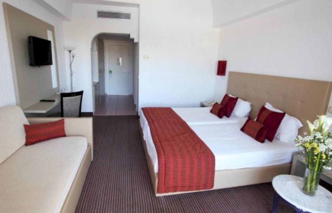 Hotelzimmer im Djerba Holiday Beach mit Doppelbett, Sofa, Fernseher und Tisch mit Blumen.