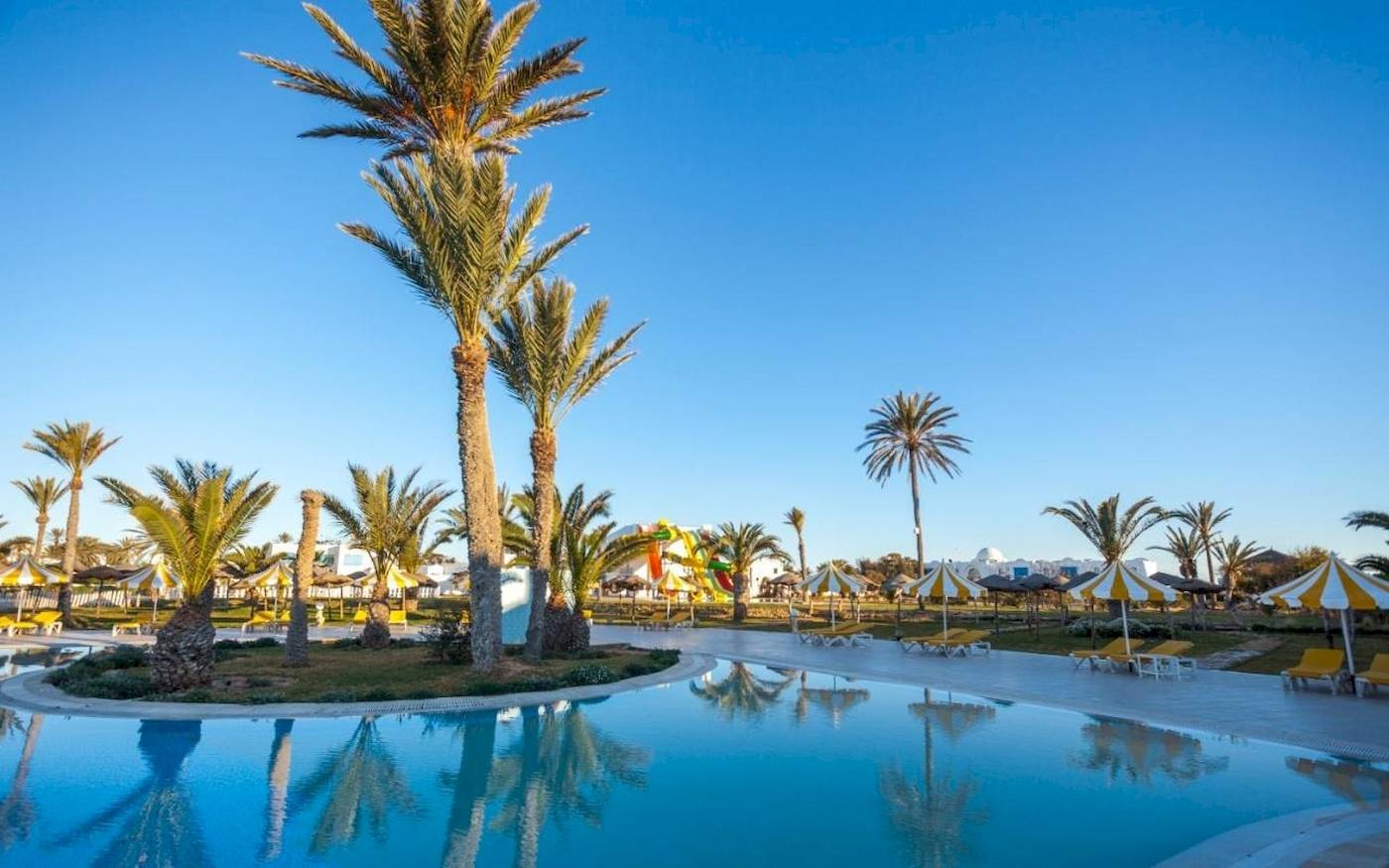 Außenpoolbereich des Djerba Holiday Beach Hotels mit Palmen, Sonnenliegen und gelben Sonnenschirmen.