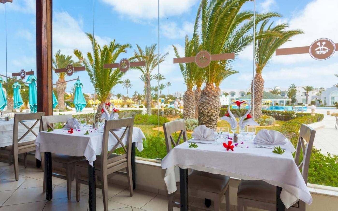 Innenansicht des Restaurants des Djerba Holiday Beach Hotels mit Tischen und Stühlen, Palmen und einem Swimmingpool im Hintergrund.