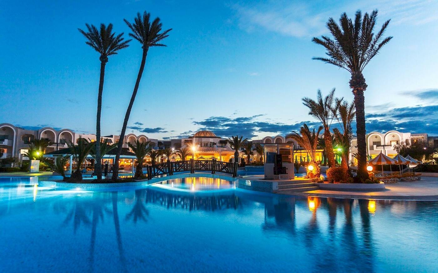 Ein Swimmingpool des Djerba Holiday Beach Hotels bei Nacht, umgeben von Palmen und Gebäuden.