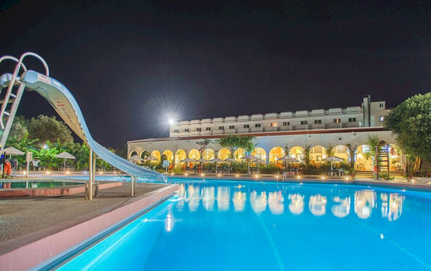 Nachtansicht des Swimmingpools im Irene Palace Beach Resort mit einer Wasserrutsche und beleuchtetem Gebäude.