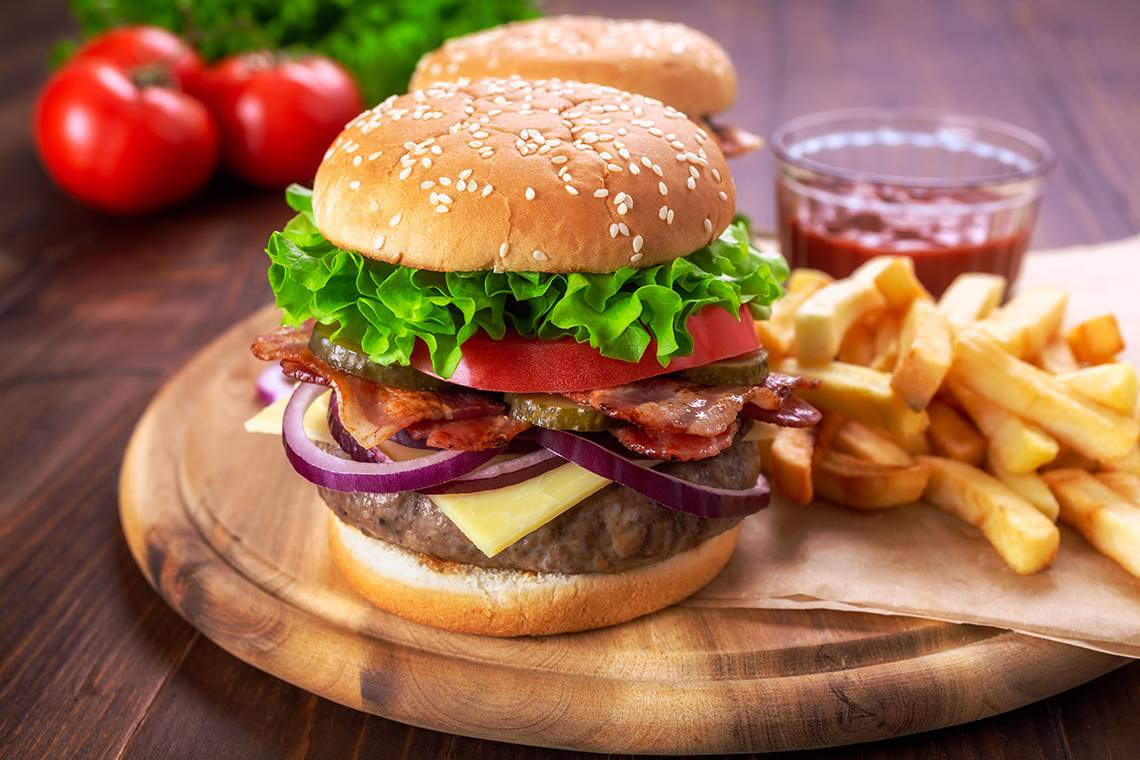 Ein Burger mit Salat, Tomaten, Zwiebeln, Schinken und Käse, serviert auf einer Holzplatte mit Pommes und Ketchup.
