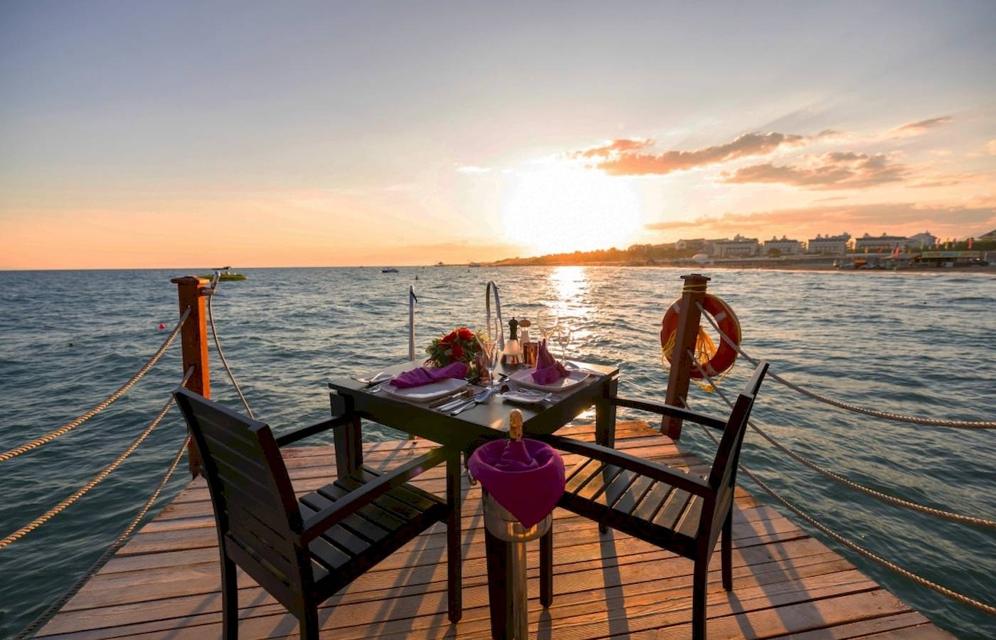 Ein Tisch auf einer Holzplattform am Wasser bei Sonnenuntergang, mit Flaschen und Blumenarrangement.
