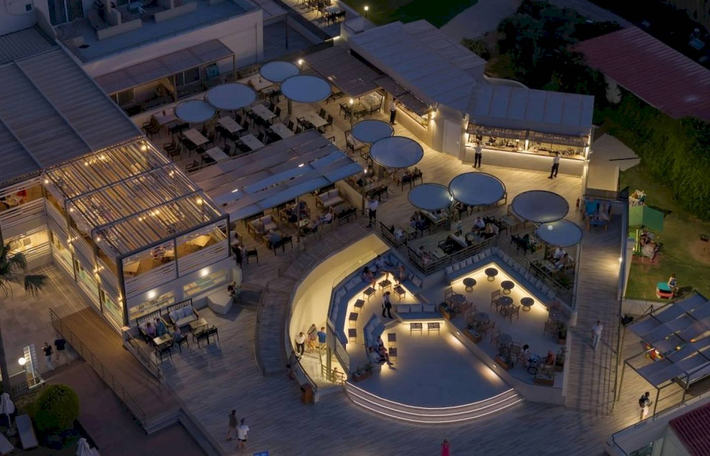 Abendluftaufnahme des Georgioupolis Resort Aquapark & SPA mit beleuchteten Terrassen und Gästen.