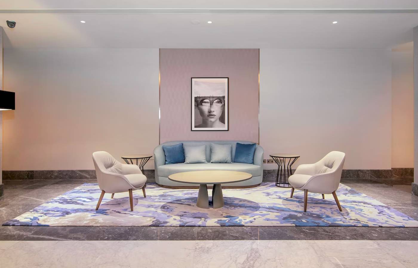 Ein modernes Hotel-Lobby mit einem blauen Sofa, weißen Sesseln und einem zentralen Tisch auf einem großen Teppich.