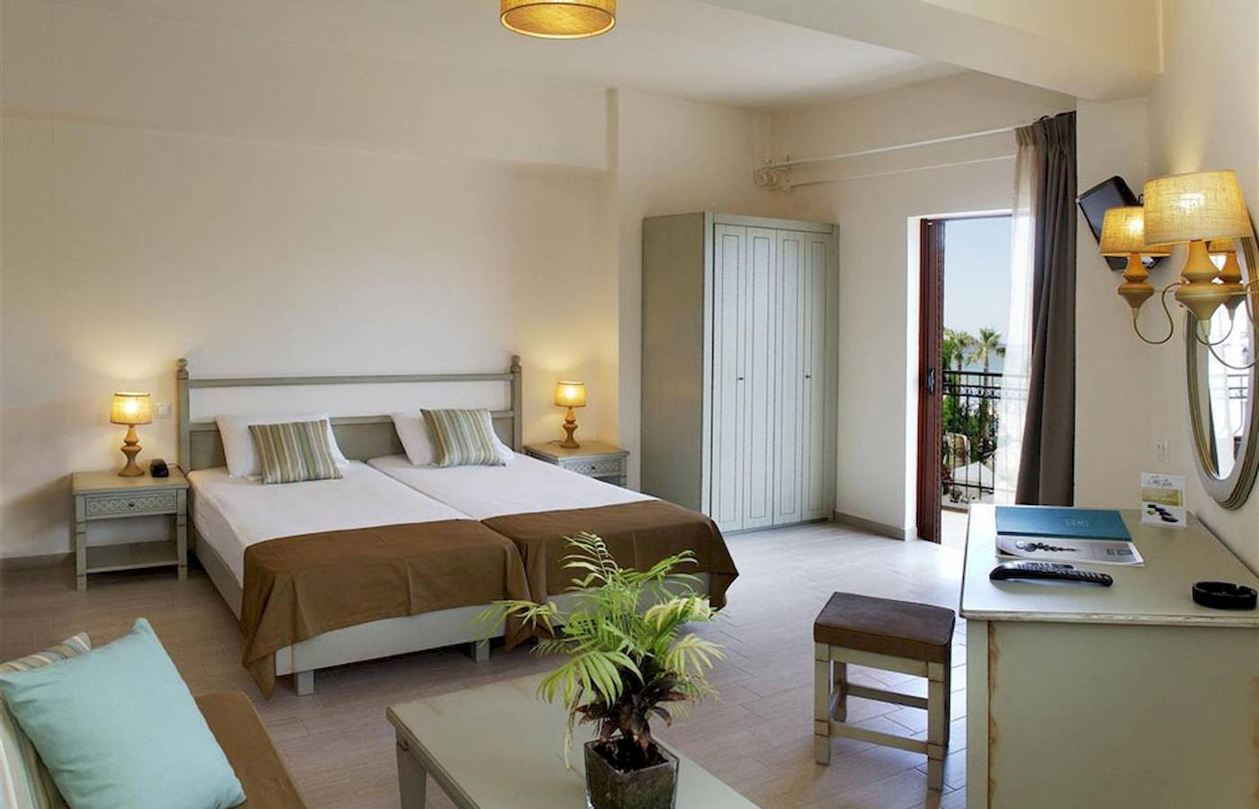 Doppelzimmer im Almyrida Beach Hotel mit zwei Betten, Nachttischen, einer Kommode, einer Lampe und einem offenen Balkon.