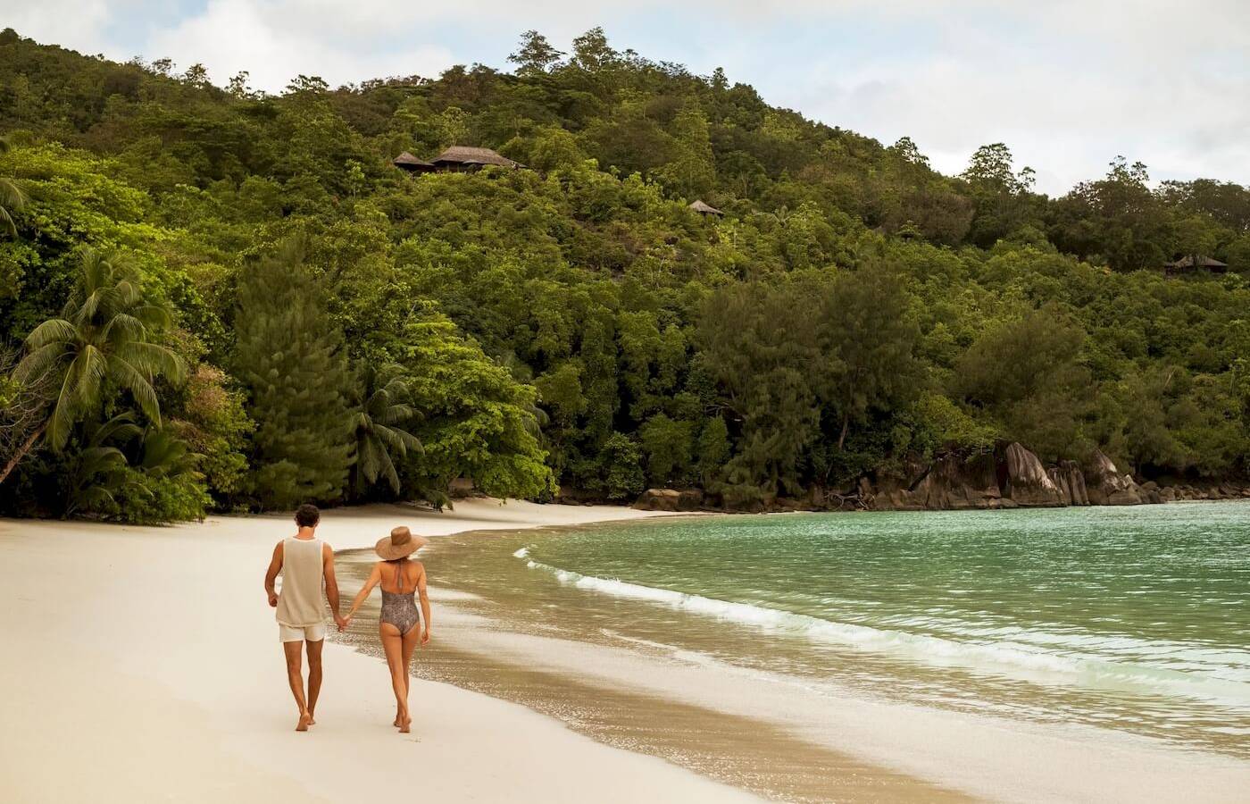 Ein Paar geht am Strand des Constance Ephelia Resorts entlang, umgeben von grüner Vegetation und kristallklarem Wasser.