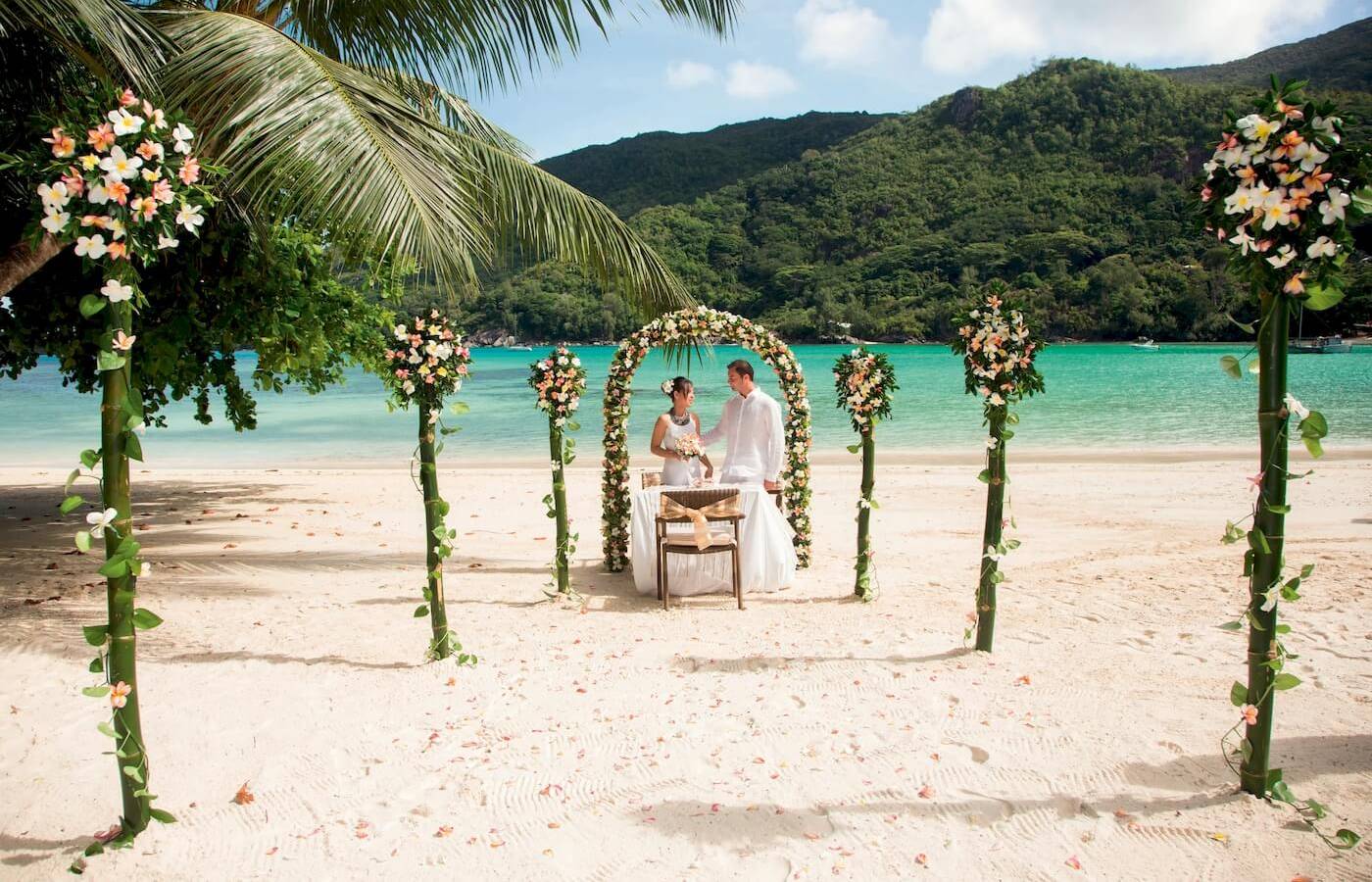 Ein Bräutigam und eine Braut stehen unter einem blumengeschmückten Bogen auf dem Strand beim Constance Ephelia Resort.