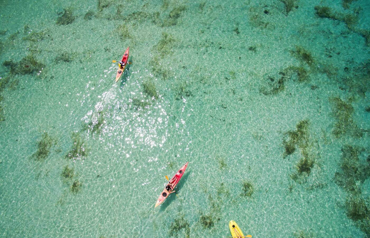 Luftaufnahme von drei Kajakfahrern im kristallklaren Wasser des Constance Ephelia Resorts.