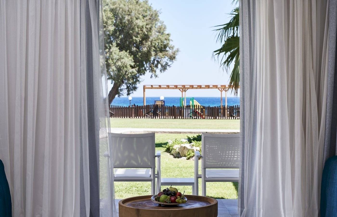 Innenansicht des Atlantica Beach Resort Kos mit Blick auf die Terrasse, auf der zwei weiße Stühle und ein runder Tisch mit Obst stehen.