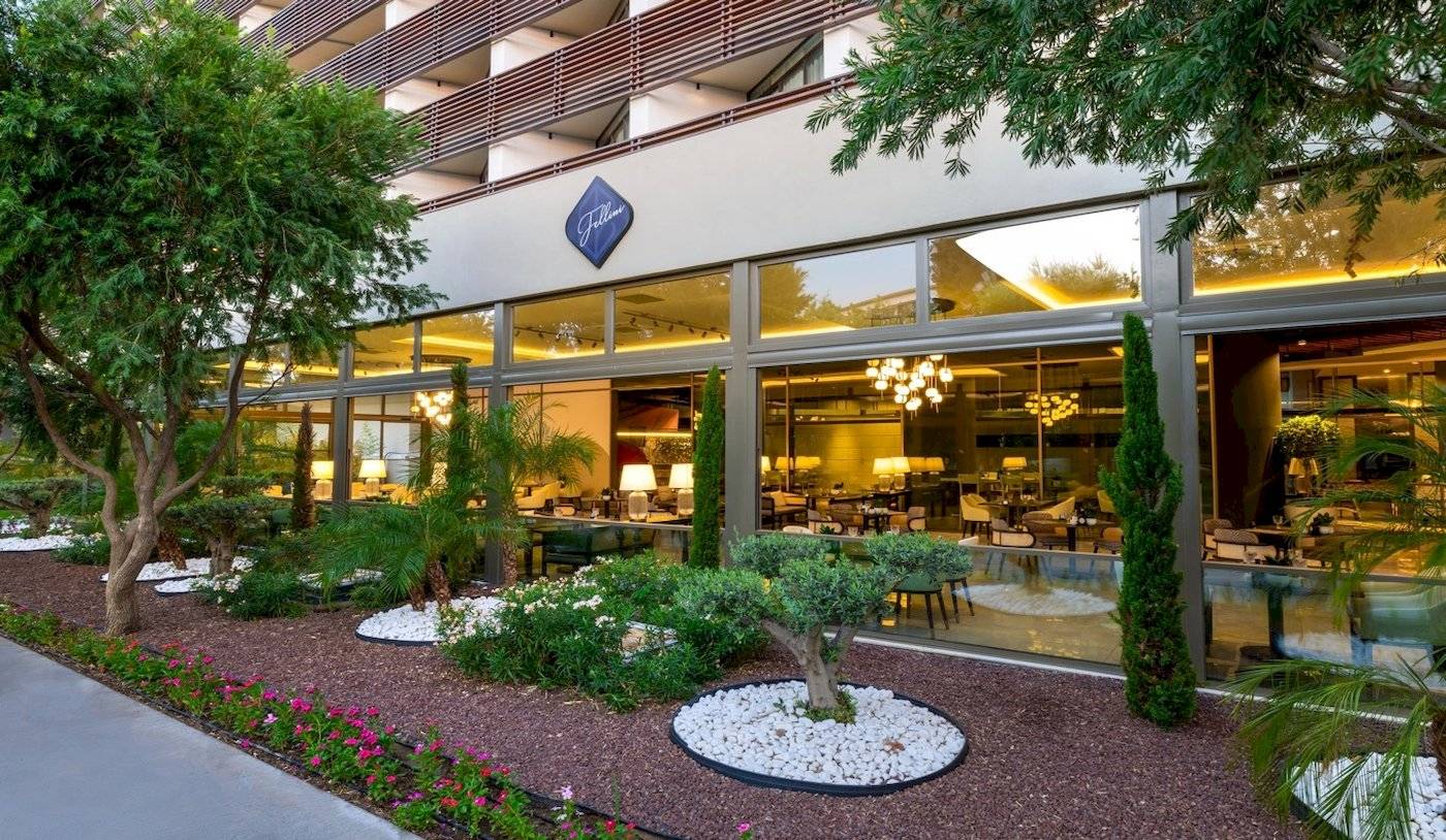 Außenansicht des Nirvana Cosmopolitan Hotels mit Garten, Bäumen und Glasfassade.