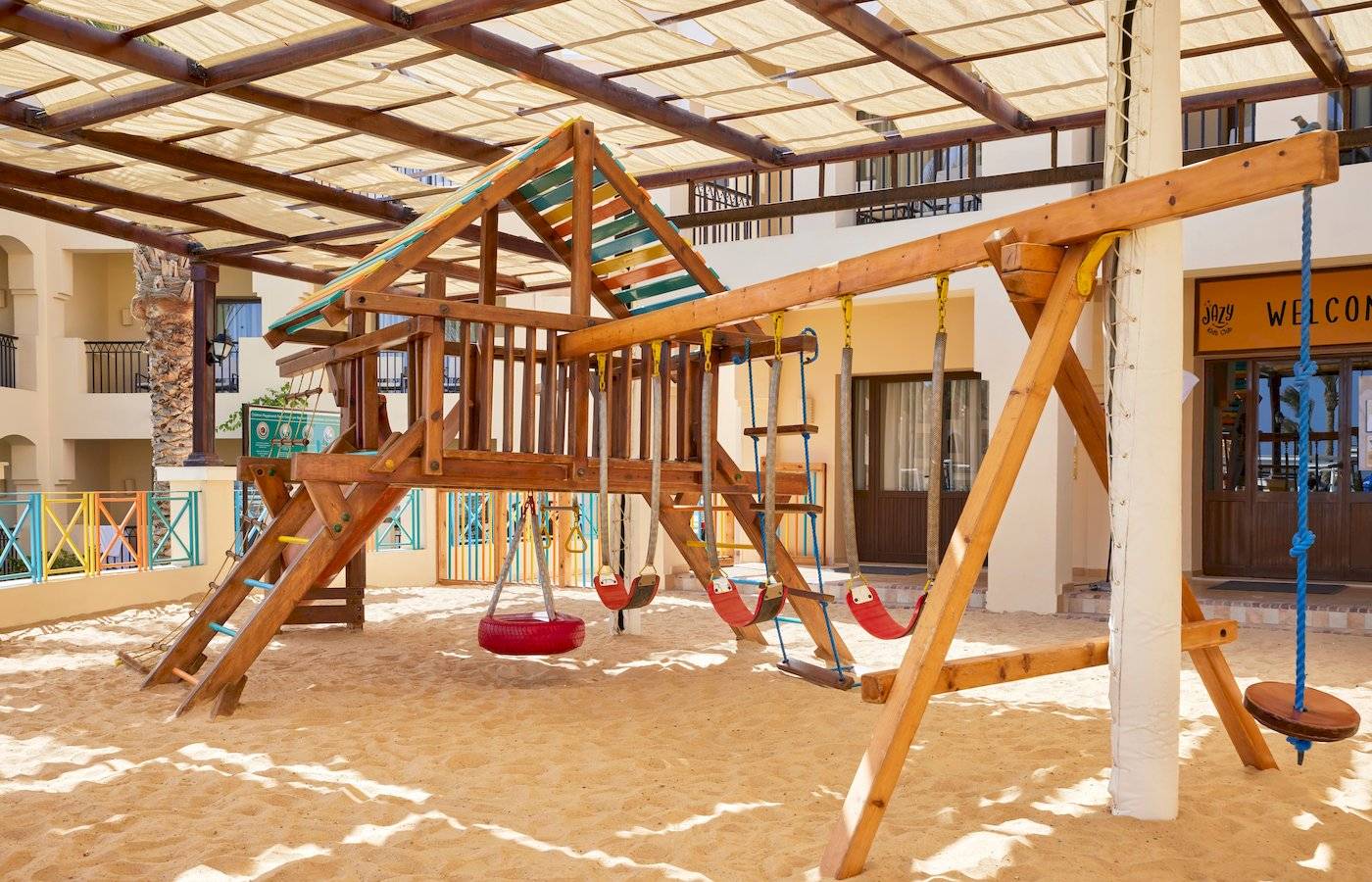 Spielplatz im Jaz Lamaya Resort mit Sandboden, Holzspielgeräten und Überdachung.