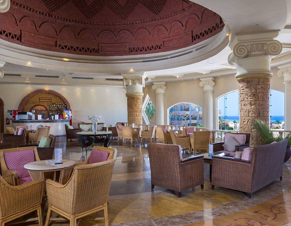 Innenbereich des Utopia Beach Club Hotels mit runden Tischen und Stühlen, einer Bar und großen Fenstern.