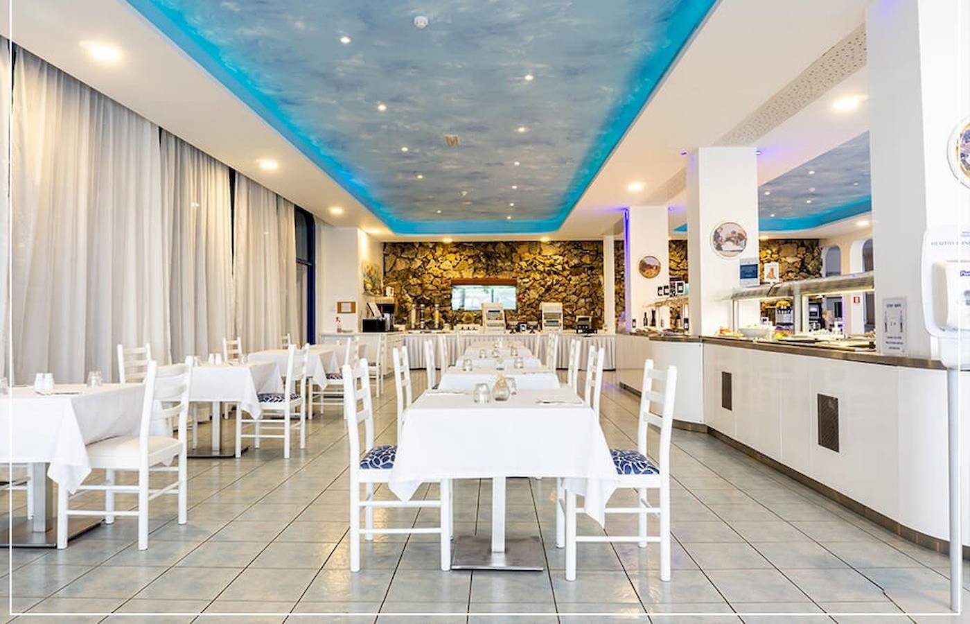 Restaurantbereich des Anonymes Strandhotel mit weißen Tischen und Stühlen, blauer Decke und Steinwand.