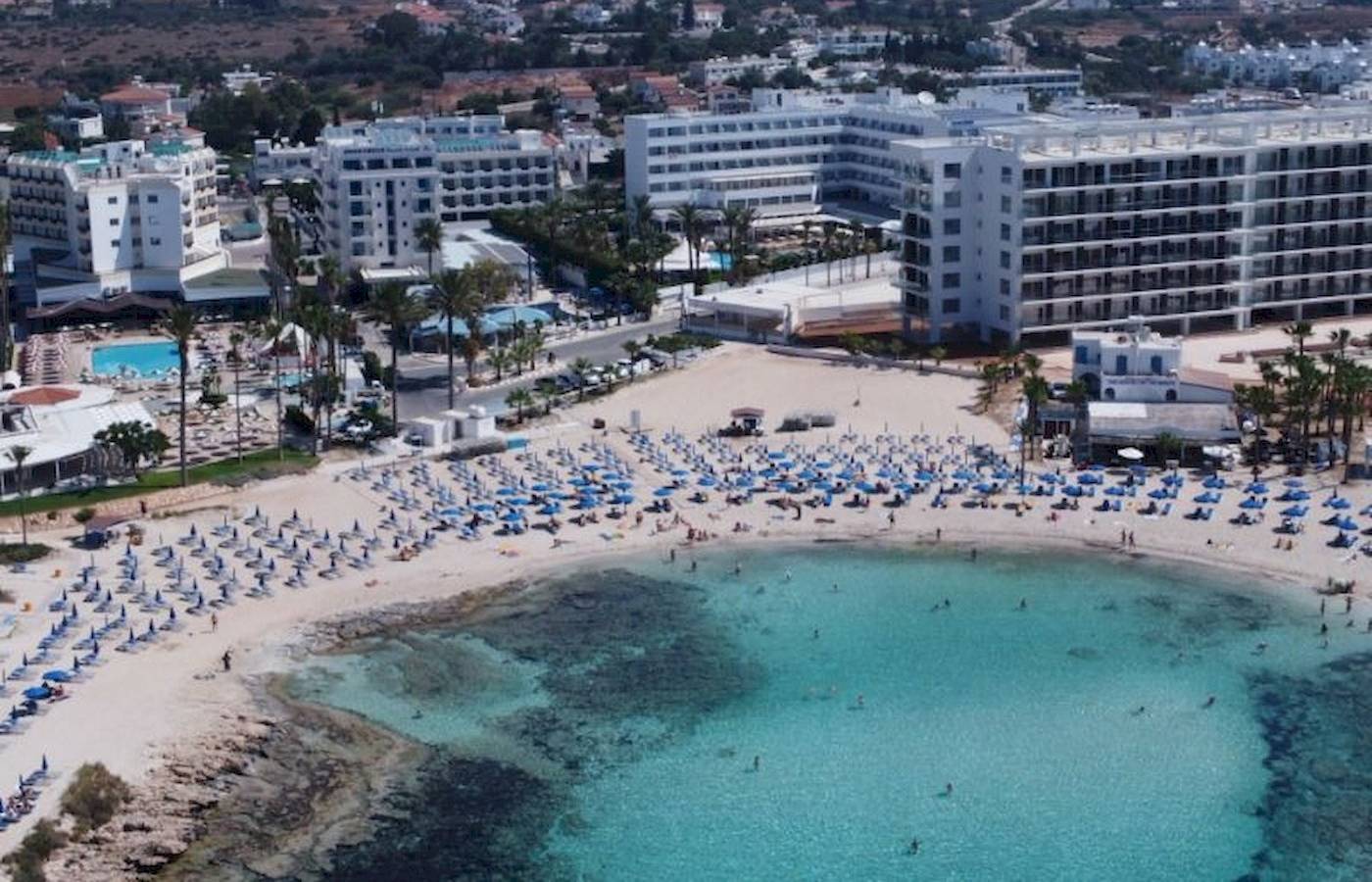 Luftaufnahme des Anonymous Beach Hotels mit vielen Gästen und blauen Sonnenschirmen am Strand.