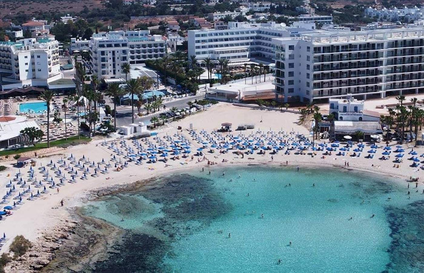 Luftaufnahme des Anonymous Beach Hotels mit vielen Gästen am Strand und blauen Sonnenschirmen.