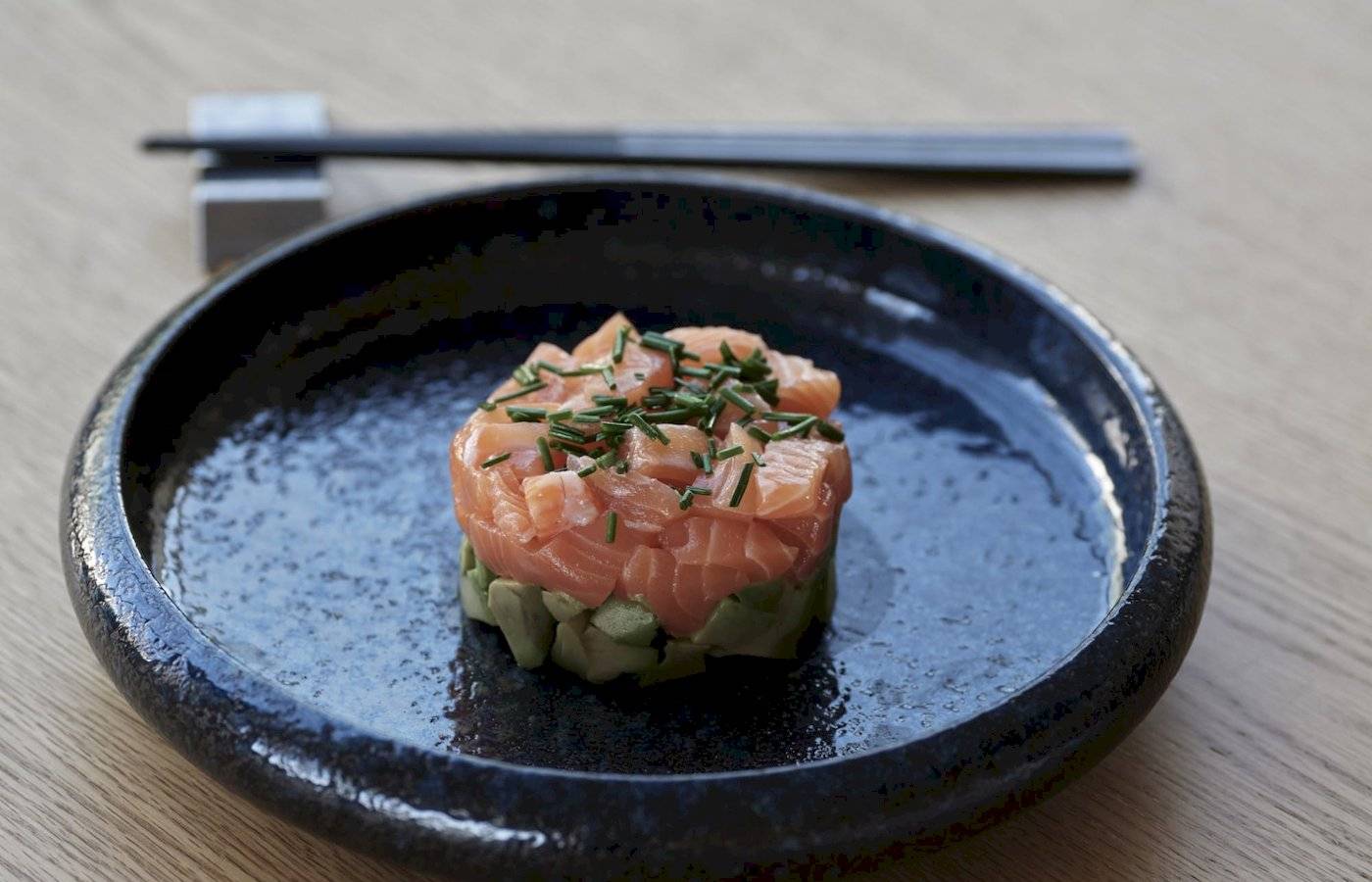 Lachs-Avocado-Tartare auf einem schwarzen Teller, auf einem Holztisch serviert, mit Essstäbchen.