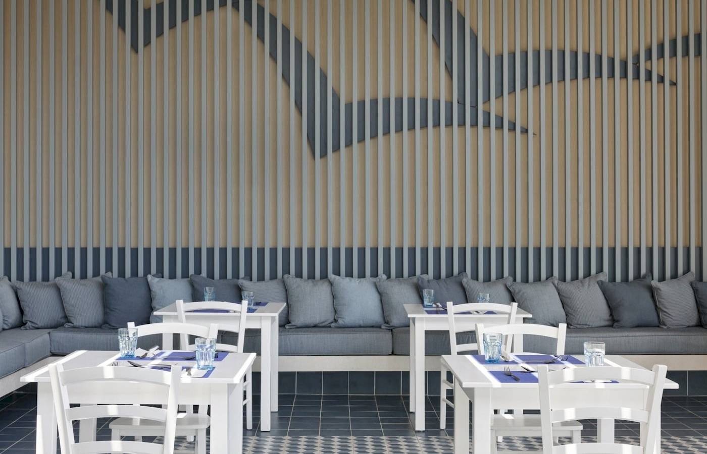 Restaurantbereich im Atlantica Kalliston Resort mit weißen Tischen und Stühlen, grauen Kissen und blauen Wanddekorationen.