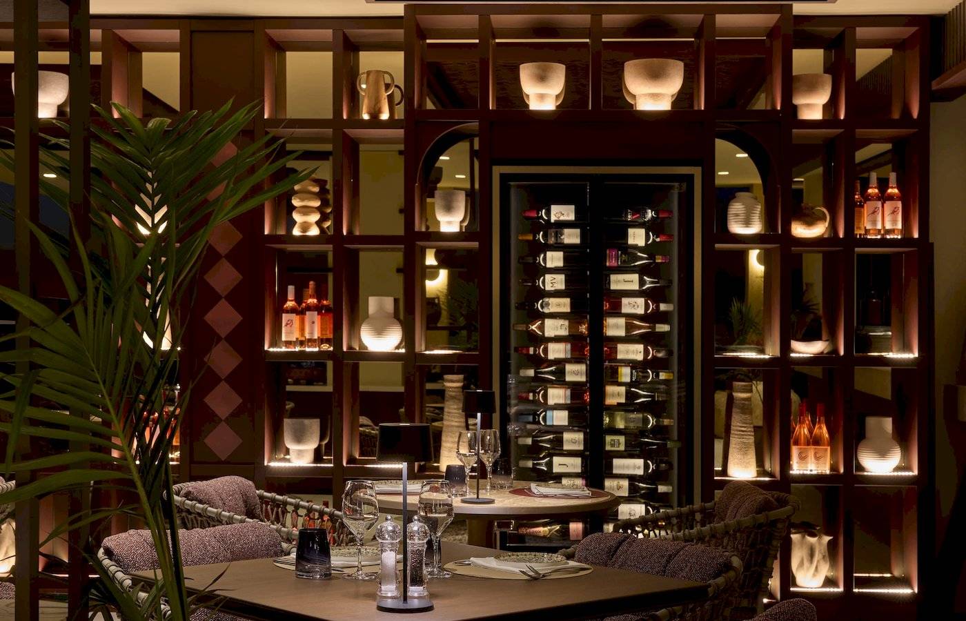 Das Interieur des Atlantica Kalliston Resorts zeigt einen eleganten Essbereich mit braunen Möbeln, Holzregalen und beleuchteten Weinflaschen.