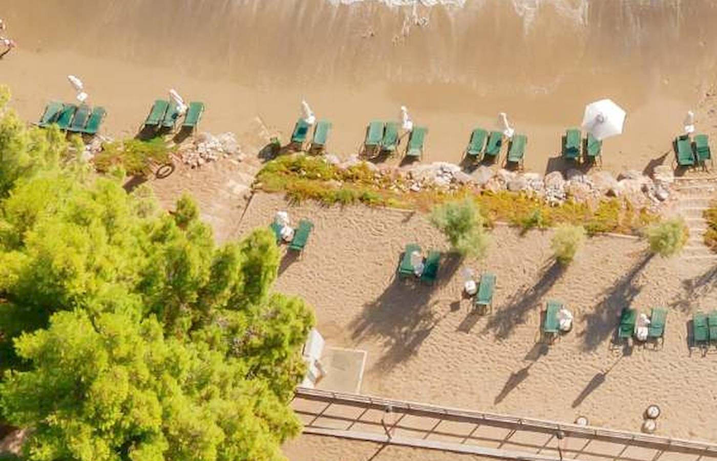 Luftaufnahme des Candia Park Village Strandes mit grünen Liegen und weißen Sonnenschirmen.