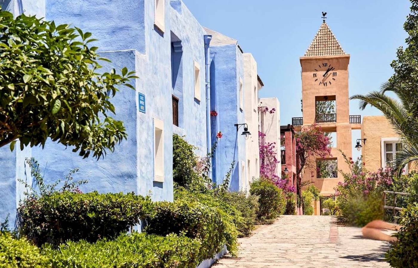 Haupteingang des Candia Park Village Hotels mit blauen Gebäuden und einer Uhrturm im Hintergrund.