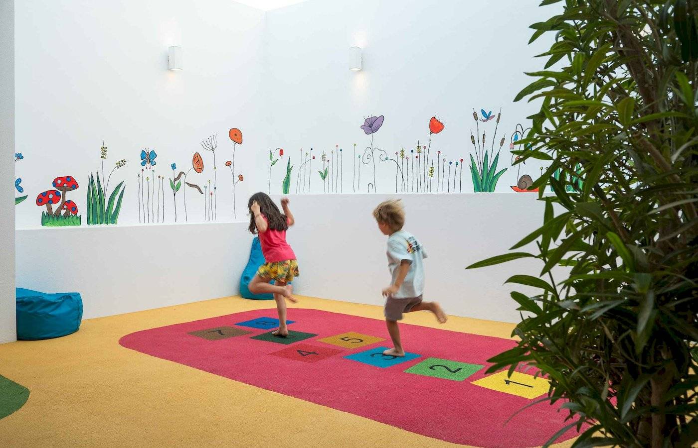 Kinder spielen auf einem roten Teppich mit nummerierten Quadraten in einem Zimmer mit Blumenwandgestaltung im Arminda Hotel & Spa.