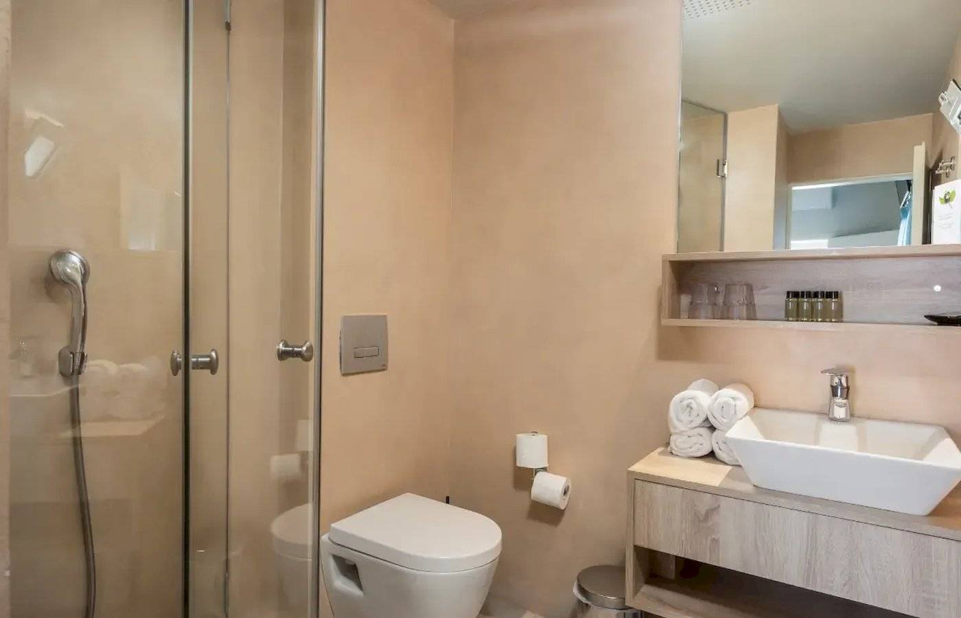 Badezimmer im Corissia Beach Hotel mit Glasdusche, Toilette, Waschbecken und Spiegel.