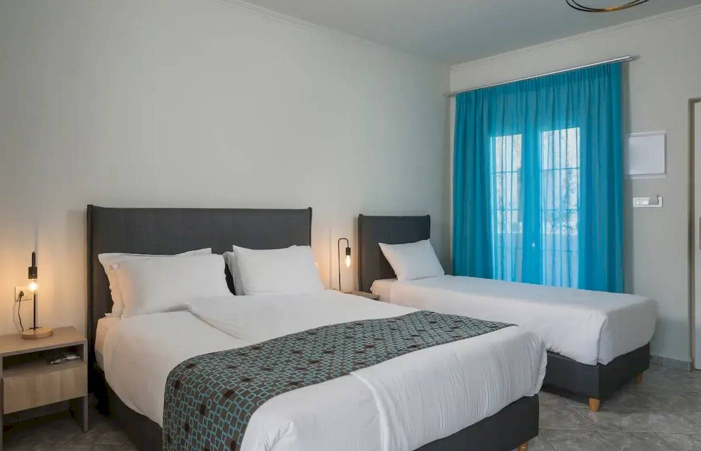 Zweibettzimmer im Corissia Beach Hotel mit weißen Wänden, zwei Betten und blauen Vorhängen.