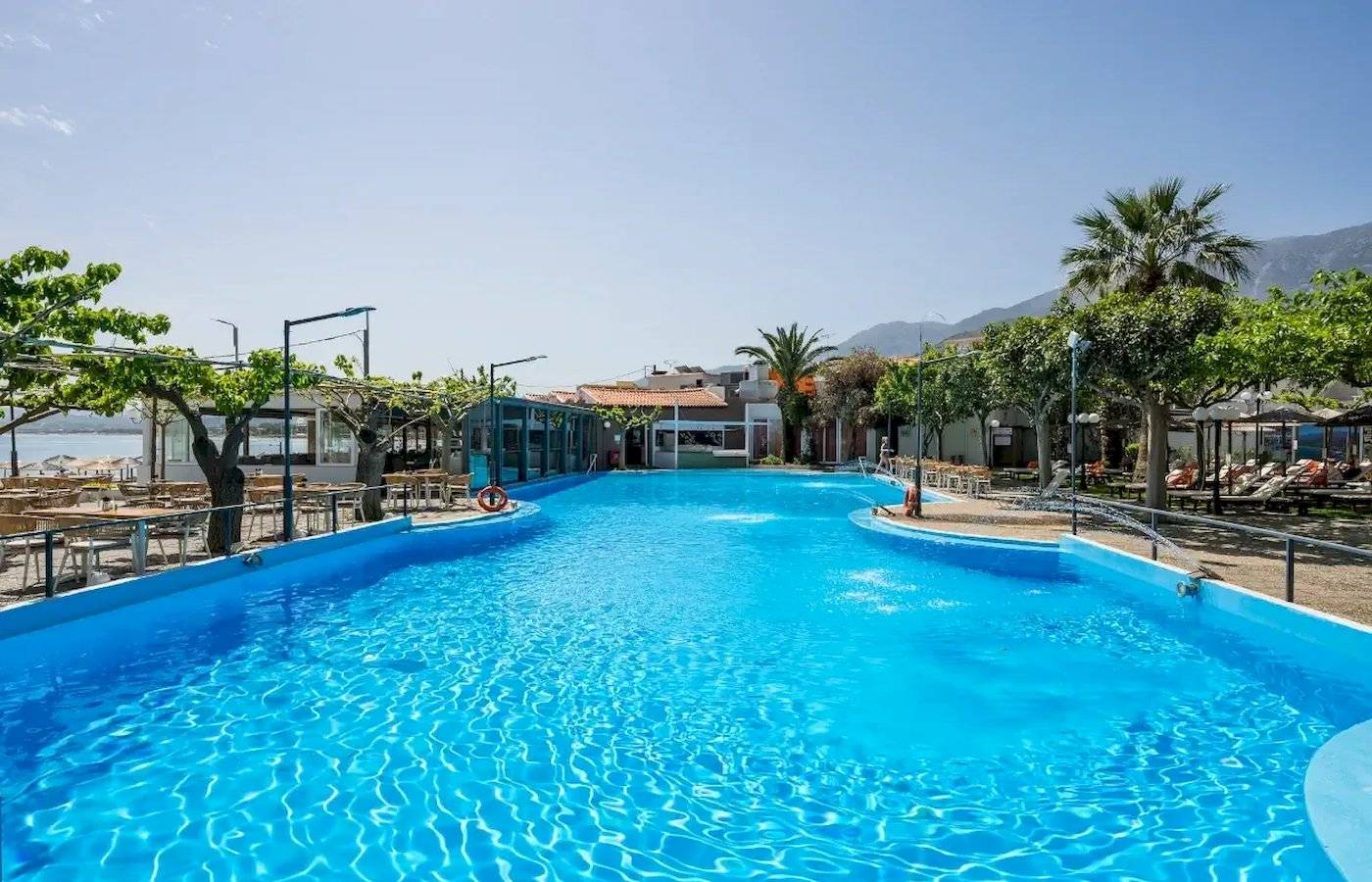 Ein großer blauer Swimmingpool im Corissia Beach Hotel mit umliegenden Bäumen und Bergen.