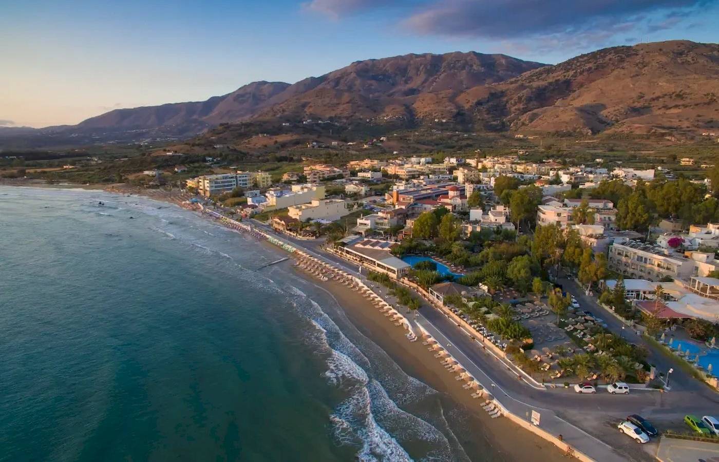 Luftaufnahme des Corissia Beach Hotels mit Strand, Gebäuden und Bergen im Hintergrund.