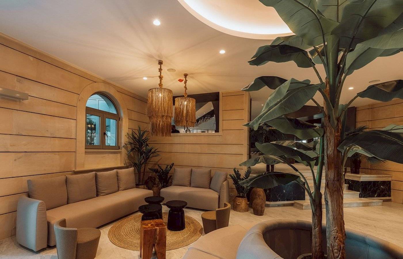 Loungebereich im Eliros Mare Hotel mit beigefarbenen Sofas, Holztischen, Pflanzen und Kronleuchtern.