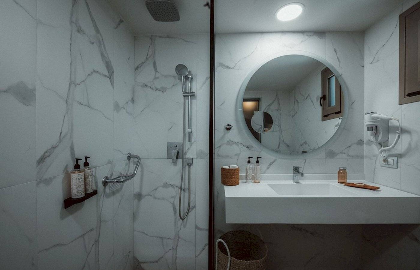 Ein Badezimmer im Eliros Mare Hotel mit einer weißen Marmorwand, einer Glasdusche, einem Spiegel und einem Waschbecken.