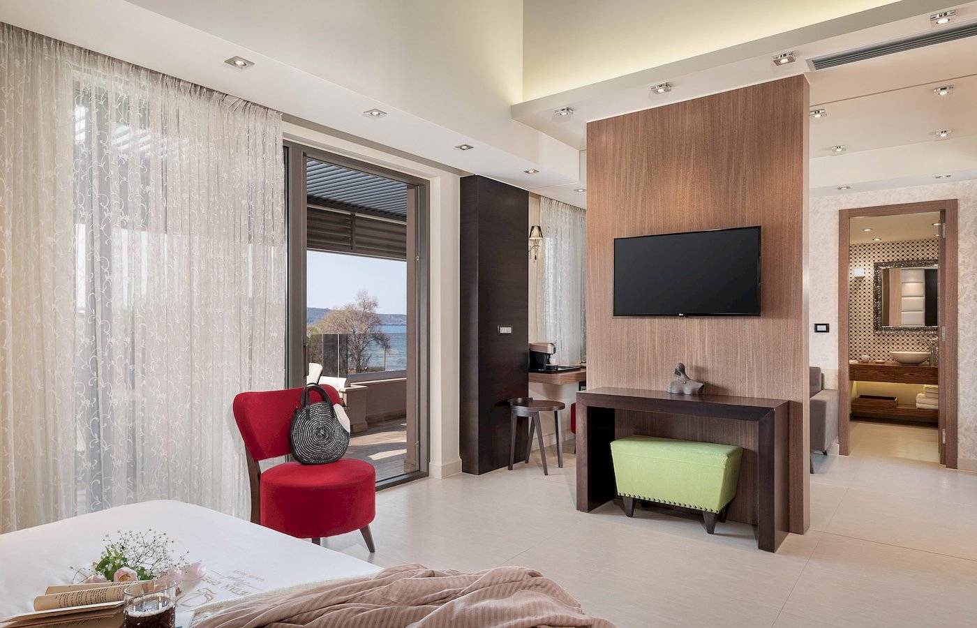 Innenansicht eines Zimmers im Cretan Dream Resort & Spa mit Bett, rotem Stuhl, Fernseher und Aussicht auf das Meer.