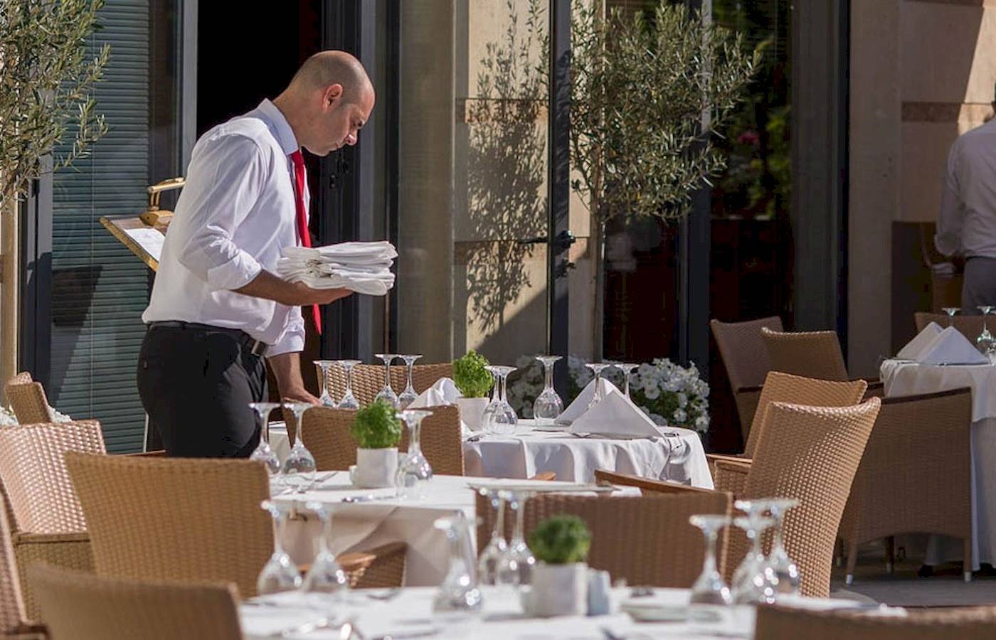 Ein Mann bereitet im Freien die Tische im Restaurant vor, wo mehrere Pflanzen und Gläser stehen.