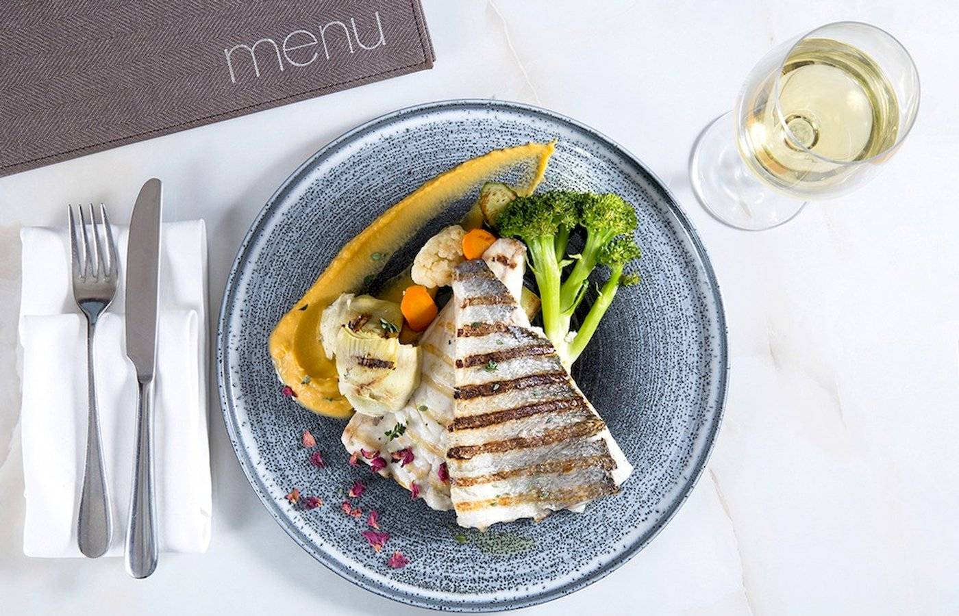 Gegrillter Fisch auf Teller mit Gemüse und Weingläsern auf weißem Tisch