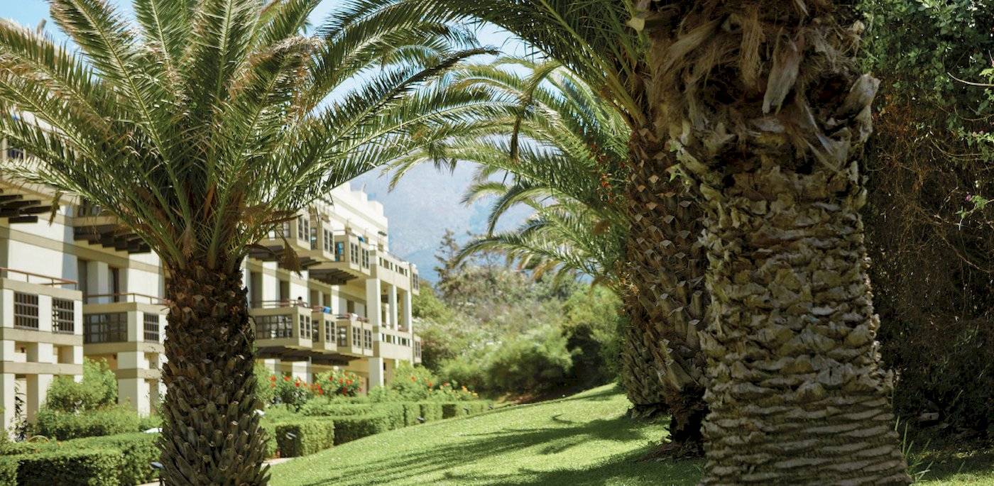 Das Grecotel Meli Palace Hotel mit Palmen und gepflegtem Rasen im Vordergrund.