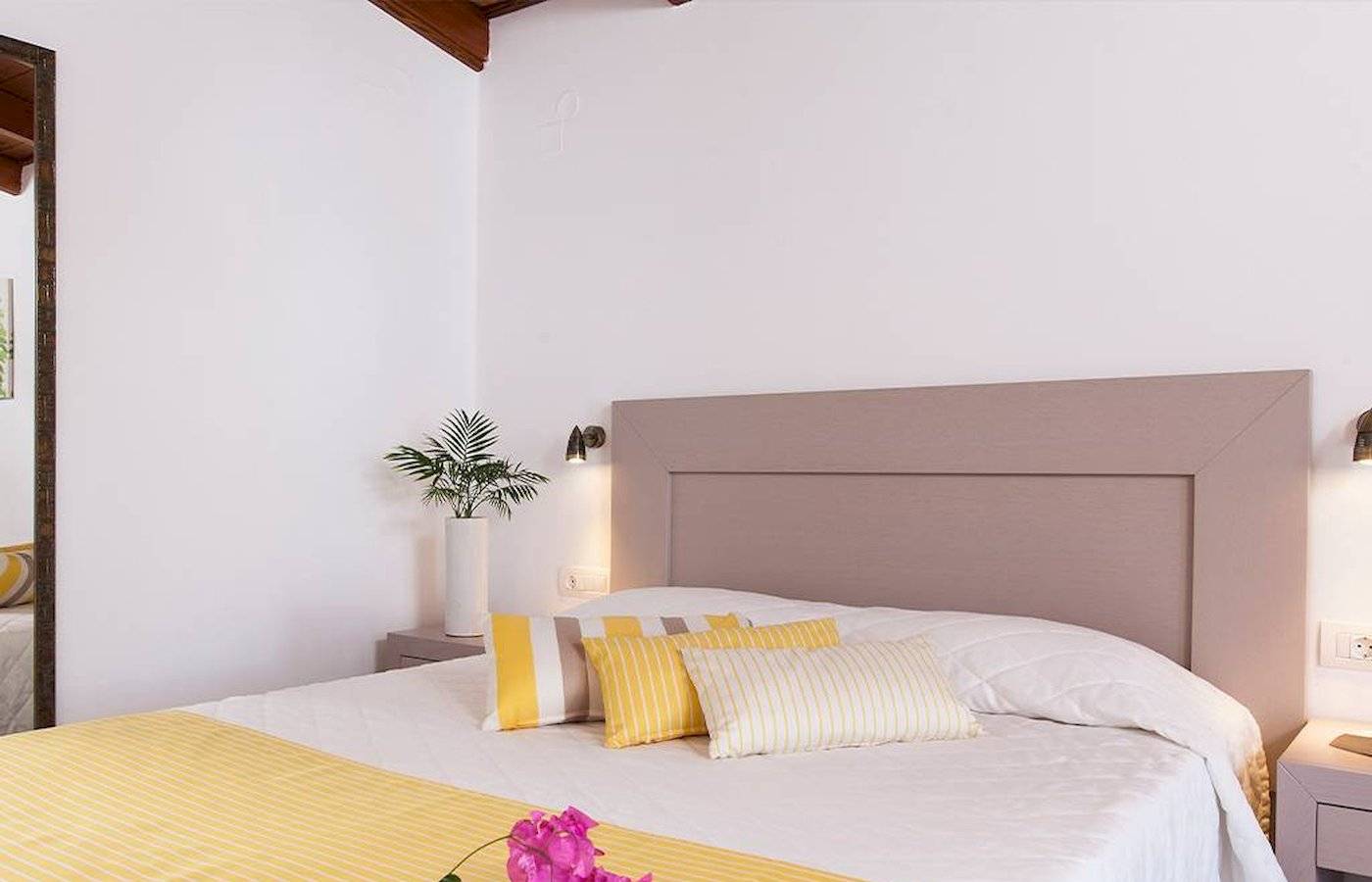 Schlafzimmer im Chrissi Amoudia Hotel mit weißen Wänden, gelben und weißen Kissen, und Blumen auf dem Bett.