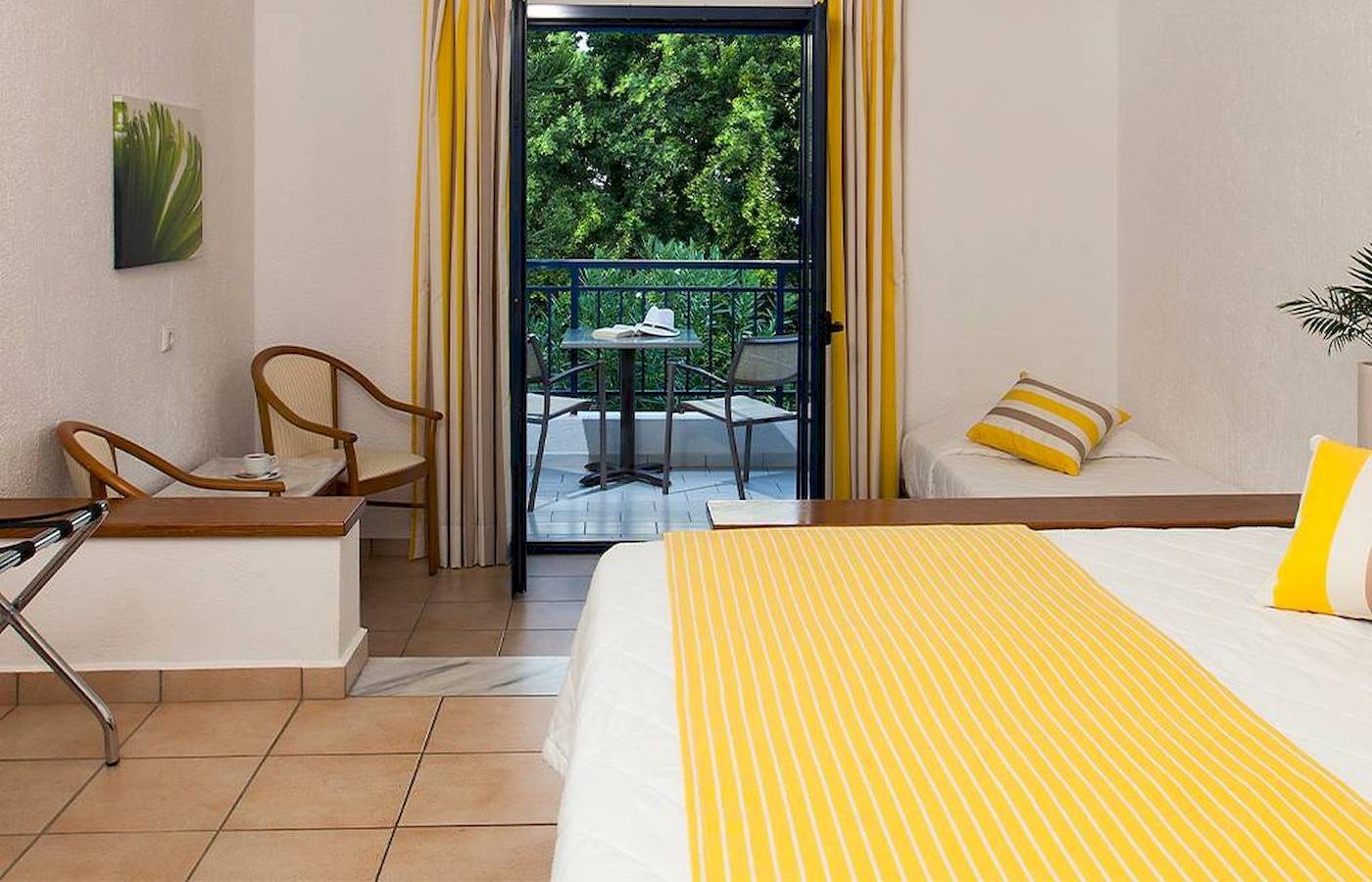 Hotelzimmer von Chrissi Amoudia mit Balkon, Bett, Tisch, Stühlen und Blick auf Bäume.