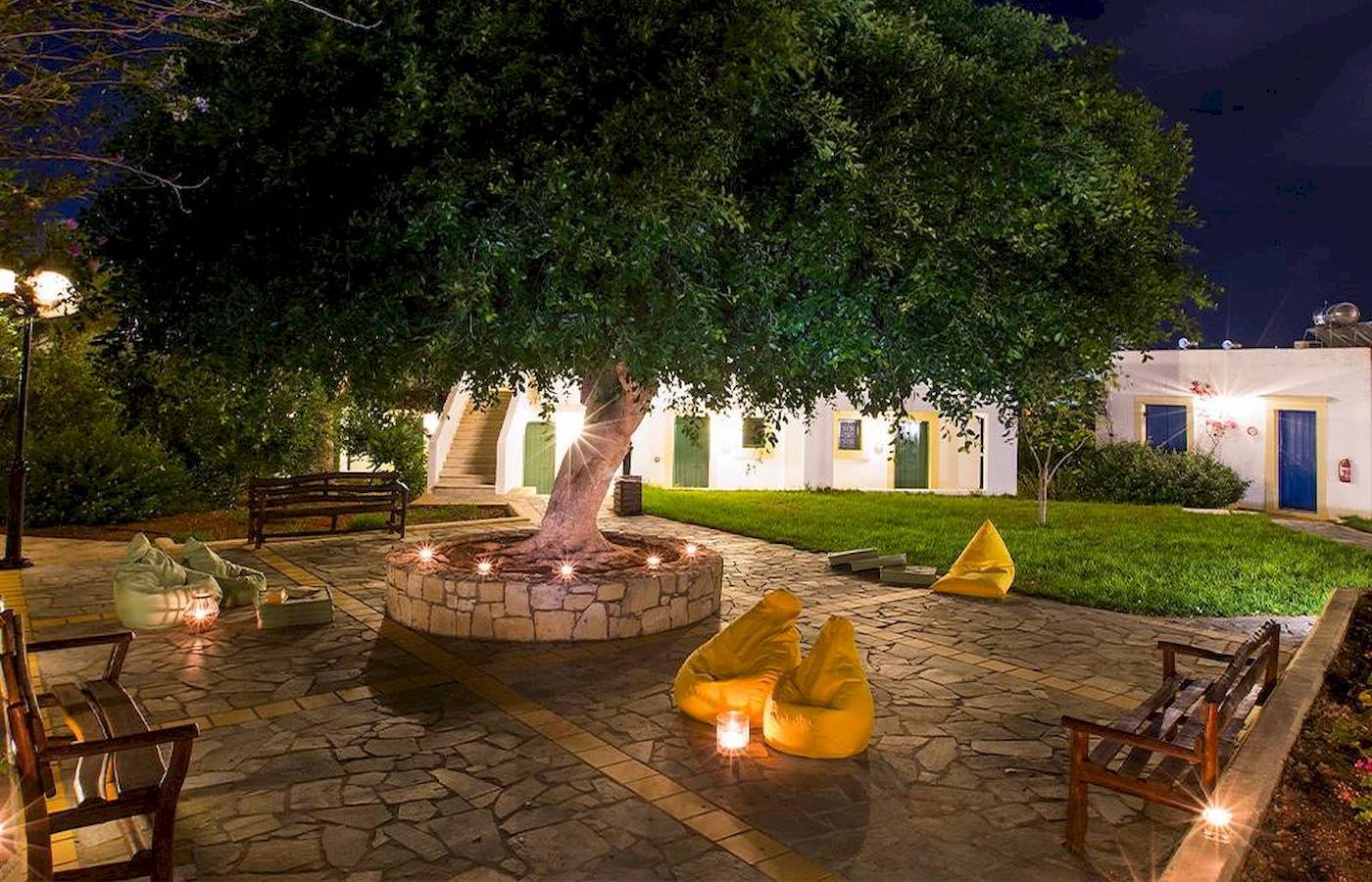 Chrissi Amoudia Hotel Atrium mit großer zentraler Baum, gelben Sitzsäcken und Nachtbeleuchtung.