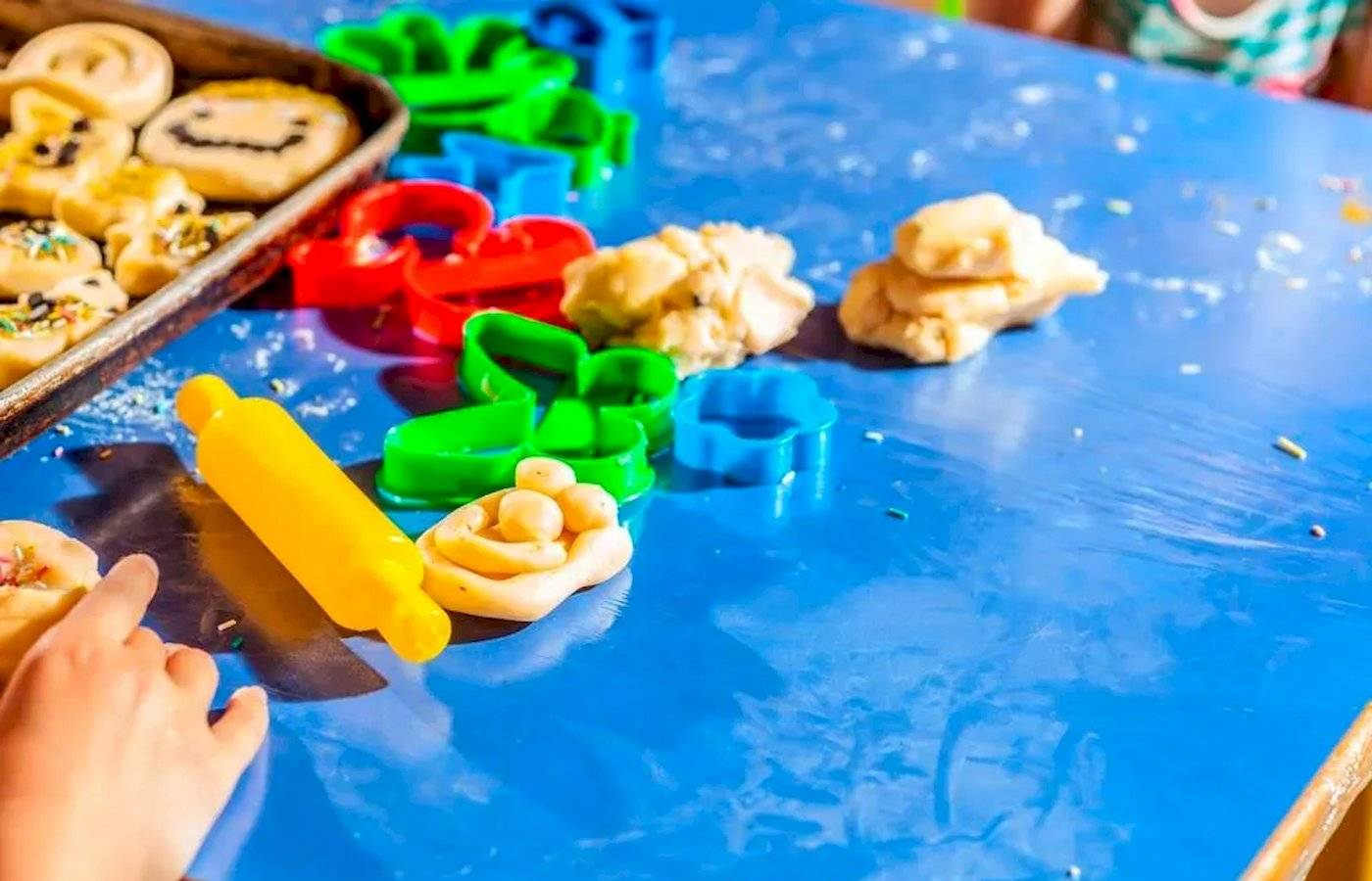 Kinder backen Kekse mit Ausstechern und einer Walze auf einem blauen Tisch.