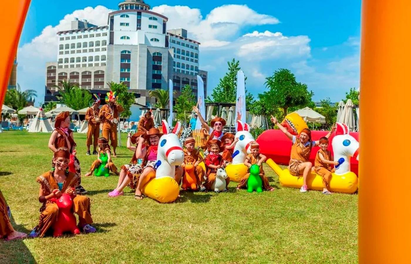 Gruppe von Kindern und Erwachsenen, die auf aufblasbaren Pferden und anderen Spielgeräten im Freien vor dem Melas Lara Hotel spielen.