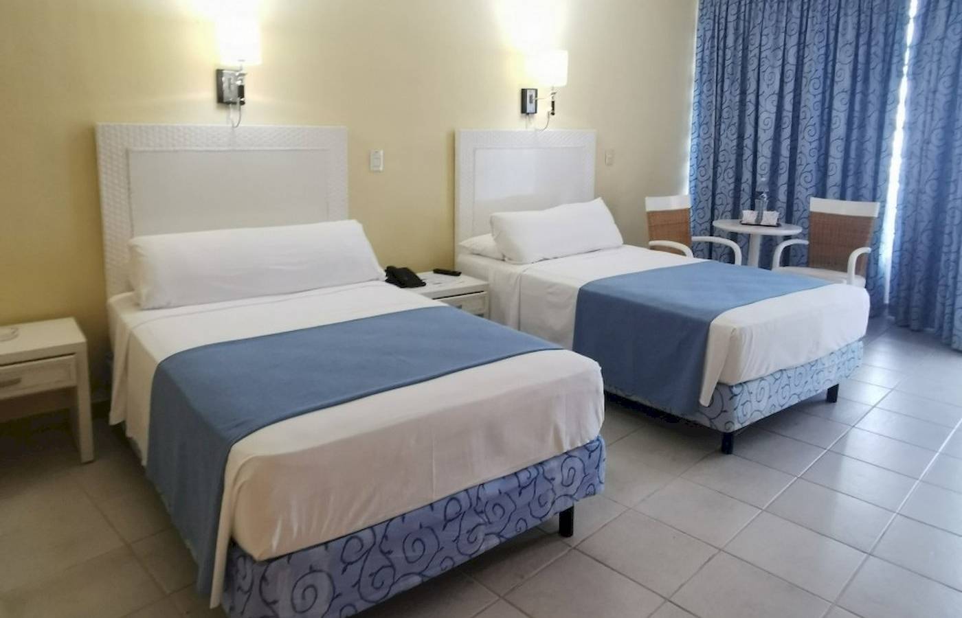 Zwei Doppelbetten im Zimmer des Muthu Playa Varadero Hotels, jedes mit weißen und blauen Laken, Nachttischen und Lampen an der Wand.