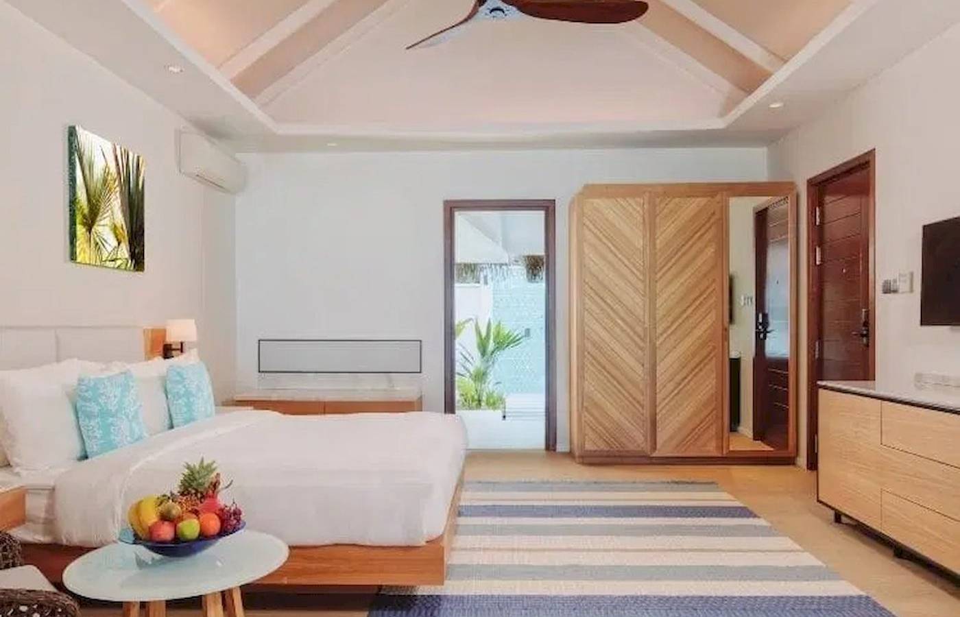 Schlafzimmer im Nova Maldives Hotel mit weißem Bett, Holzmöbeln und blau-weiß gestreiftem Teppich.