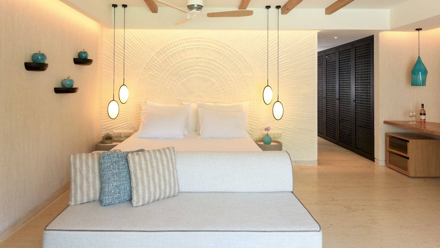 Schlafzimmer im Mitsis Selection Rinela Hotel mit Doppelbett, Couch, Deckenventilator und Wandleuchten.