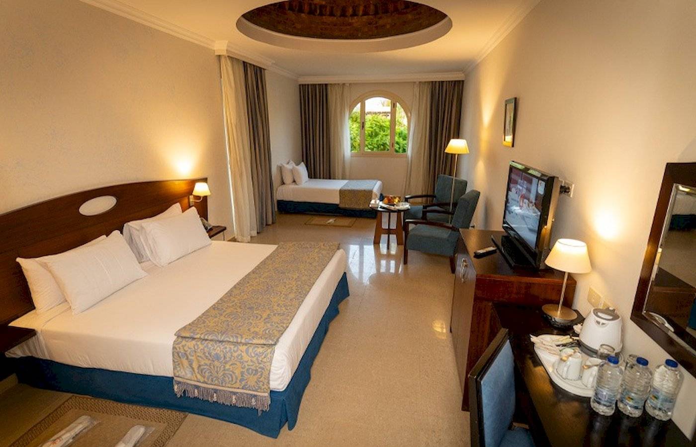 Zimmer im Stella Gardens Resort & Spa Makadi Bay mit Doppelbett, Nachttisch, Fernseher, Sessel und Minibar.