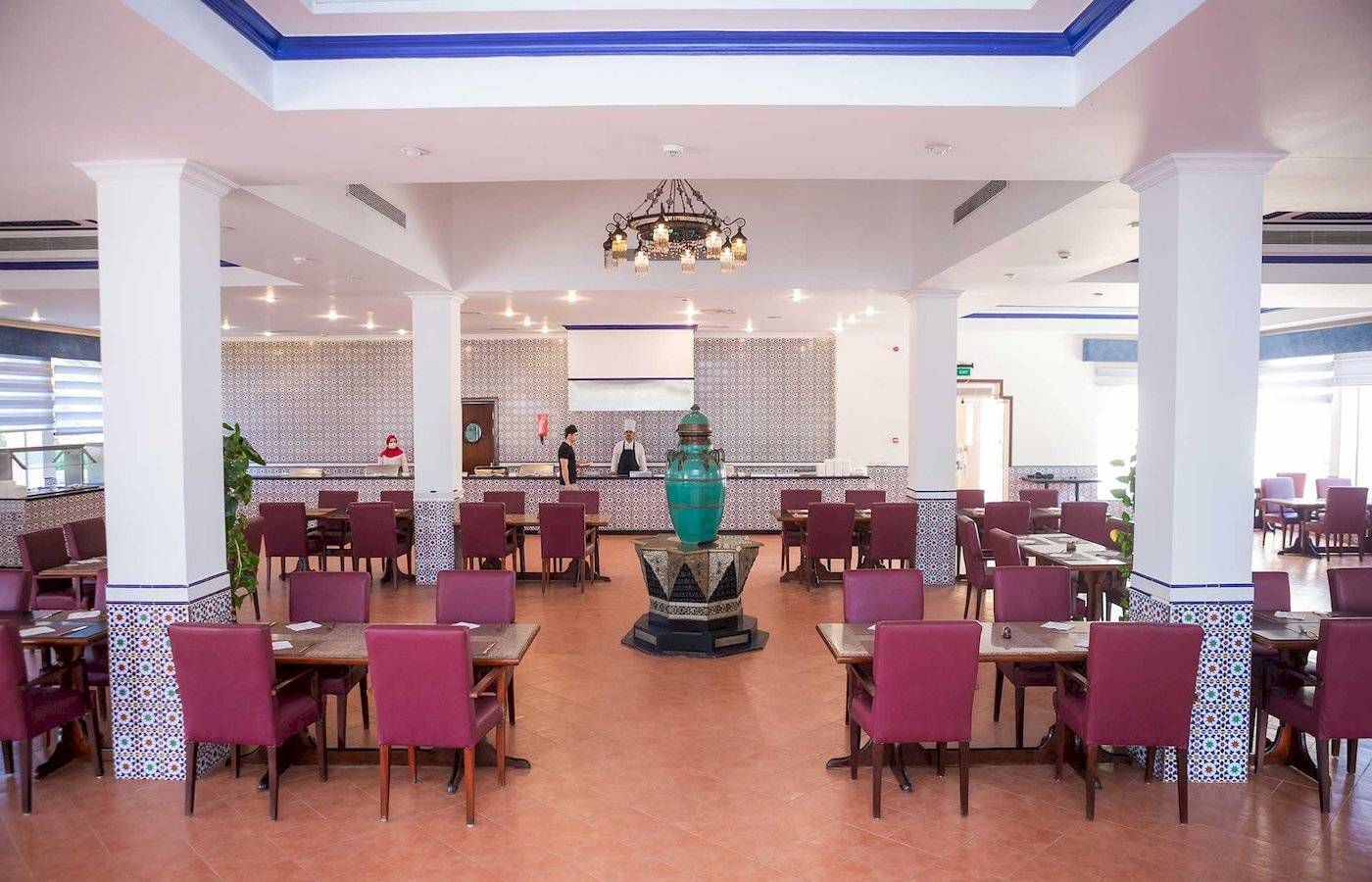 Innenbereich des Stella Gardens Resort & Spa Makadi Bay mit geometrischen Wandverkleidungen, runden Tischen, roten Stühlen, grünem Brunnen und hängender Lampe.