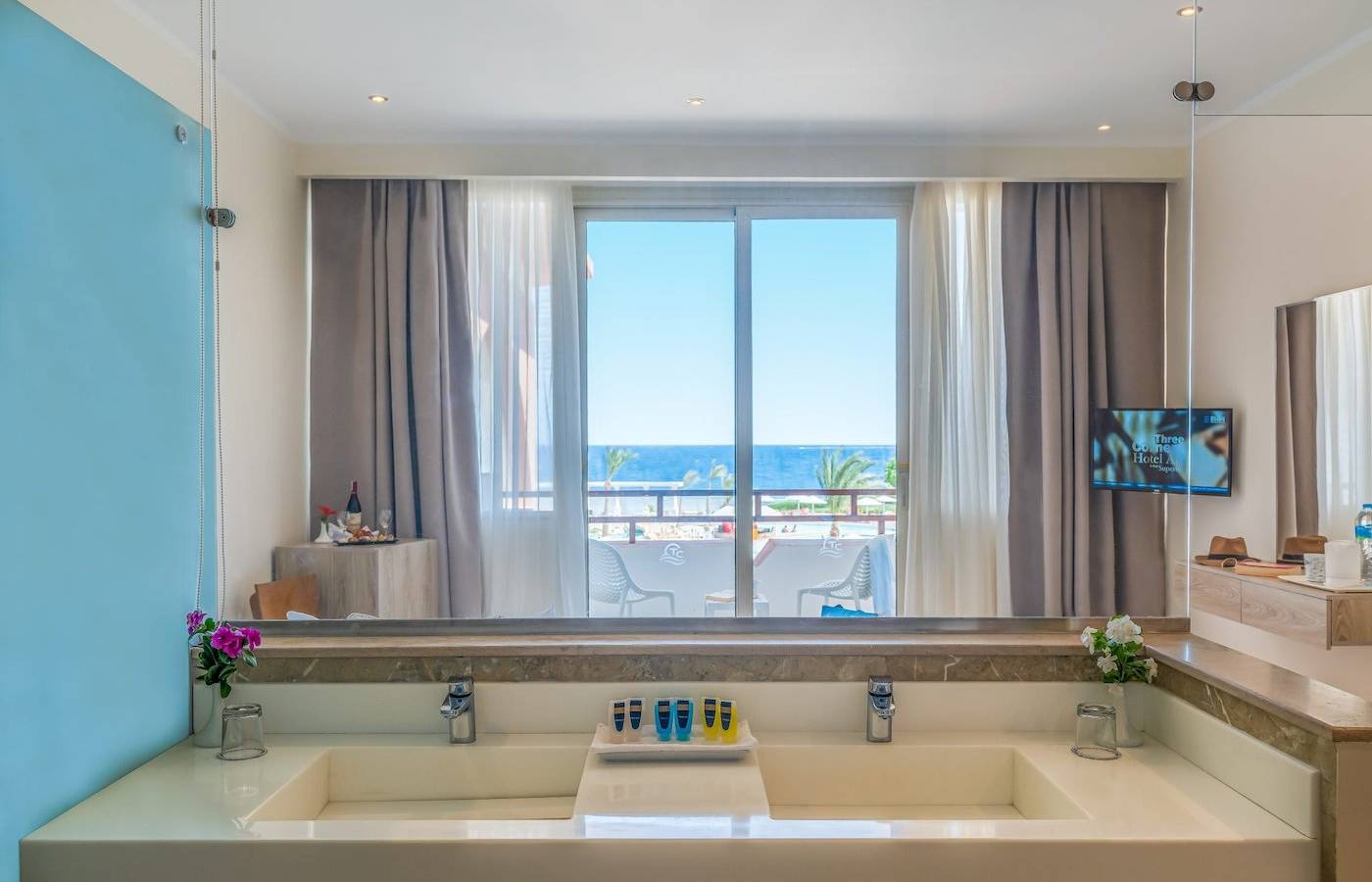 Badezimmer-Ansicht im Three Corners Equinox Beach Resort mit großem Fenster, das auf den Ozean und eine Terrasse mit Stühlen hinausblickt.