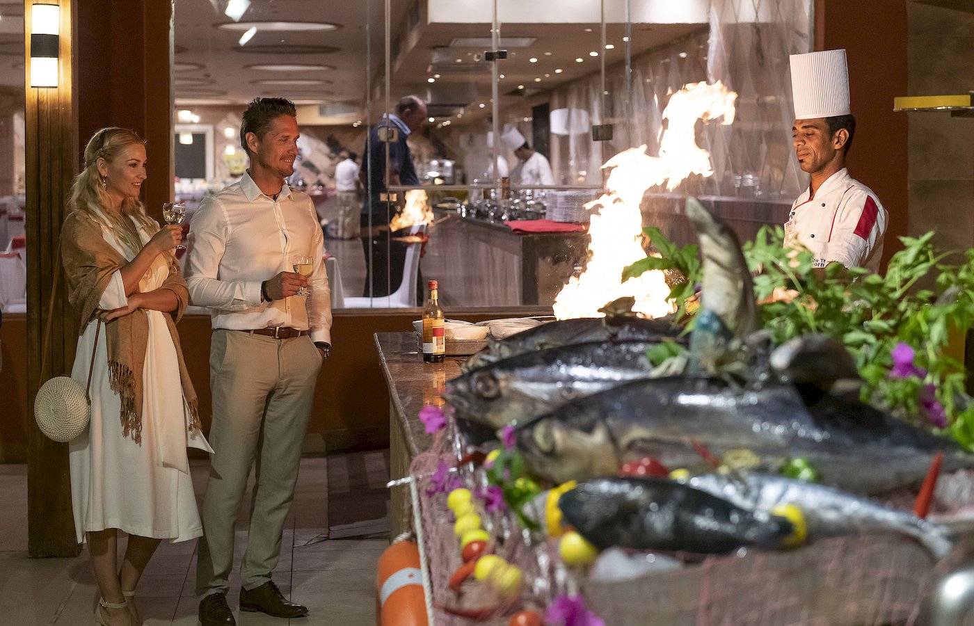 Zwei Gäste im Three Corners Happy Life Beach Resort halten Gläser mit Wein, während ein Koch ein Gericht anzündet.