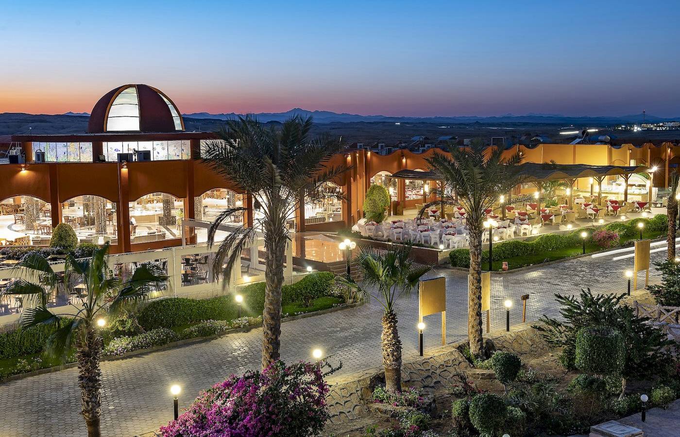 Abendansicht des Three Corners Happy Life Beach Resorts mit beleuchteten Palmen und Gebäuden.